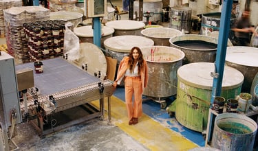 A woman in a coral suit standing in a professional paint factory with industrial mixing vats and conveyors.