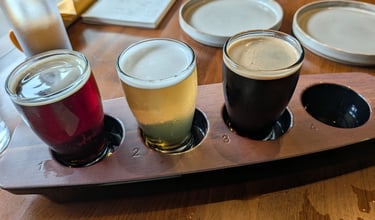 A craft beer flight with red, blonde, and dark stout samples on a wooden tasting paddle.