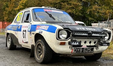 White Ford Escort MK1 rally car with sponsor decals and auxiliary light pods parked on a gravel track.