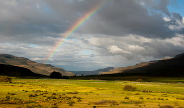 Rainbow across a cloudy sky with some fields in the foreground and a circular mountain range as backdrop