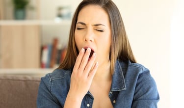 A tired young woman yawning and covering her mouth with her hand while sitting on a sofa.