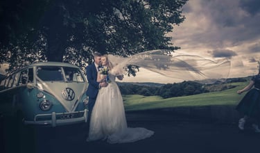 Bride and groom embrace by a vintage blue VW camper van with a flowing wedding veil in the wind.