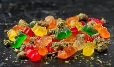 A colorful pile of THC gummy bears and dried cannabis flower buds on a dark textured surface.
