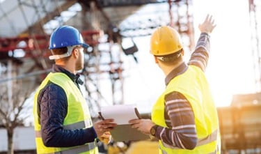 Two construction engineers wearing hard hats and safety vests inspect a building site.