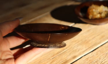 A person holding a small, polished coconut shell bowl on a rustic wooden table with natural sunlight.