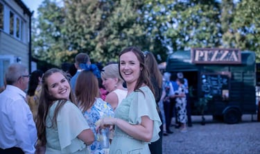 Smiling bridesmaids at an outdoor wedding reception with a horsebox pizza catering van.