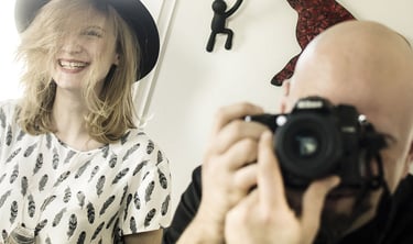 A photographer, Séb taking a picture of a smiling woman wearing a black hat and feather print shirt.