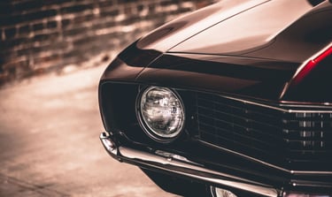 Close-up of a classic black muscle car headlight and chrome bumper with a vintage brick background.