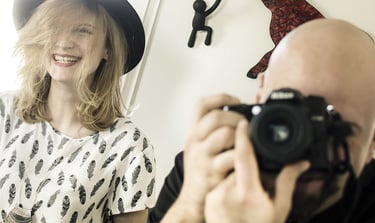 A photographer, Séb taking a picture of a smiling woman wearing a black hat and feather print shirt.