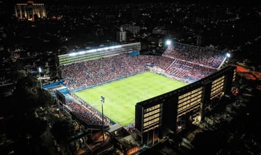 Vista panorâmica interna do Estádio Gran Parque Central com arquibancadas em azul, branco e vermelho sob luz natural.