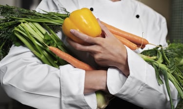 A professional chef in a white uniform holding fresh carrots and a yellow bell pepper.