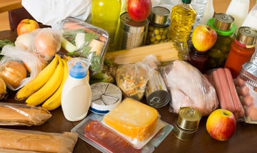 Variety of groceries including fresh fruit, vegetables, meat, cheese, and canned goods on a wooden table.