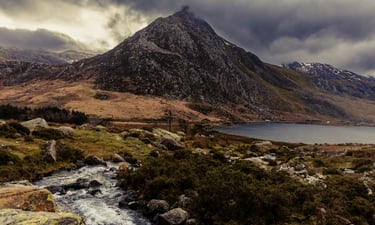 Dramatic mountain peak overlooking a scenic lake and rocky stream in Snowdonia, Wales.