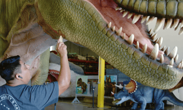An artist carefully painting a large, realistic animatronic Tyrannosaurus Rex dinosaur model in a workshop.