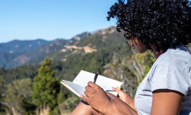 A woman with curly hair writes in a journal while sitting outdoors with a scenic mountain view.