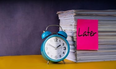 A blue alarm clock next to a stack of paperwork with a pink sticky note that says Later, symbolizing procrastination.