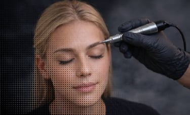 Professional technician performing a permanent makeup eyebrow tattoo procedure on a blonde woman.