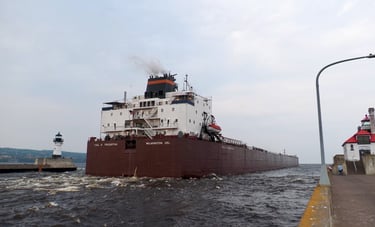 The stern of a huge red ship passing between 2 lighthouses at the entrance of the Duluth Ship Canal.