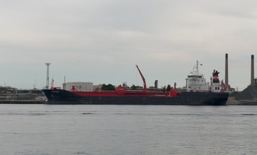A blue hulled tanker ship with a white superstructure and red deck, docked at a refinery.