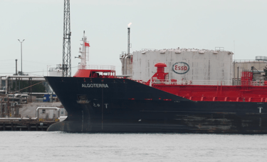 The blue bow and red deck of a tanker ship in front of large oil tanks at a refinery.