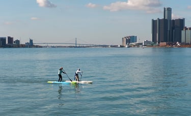 2 men paddleboarding with the Ambassador Bridge and Detroit and Windsor skylines in the background.