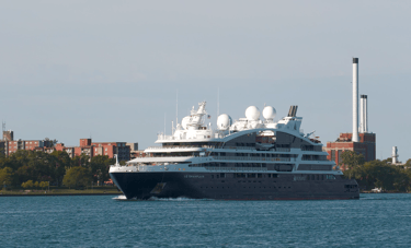 A blue and white cruise ship passing. Apartment buildings and some smokestacks in the background.