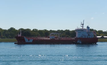 Left side view of a red tanker ship with a white superstructure and 2 white stripes near the bow.