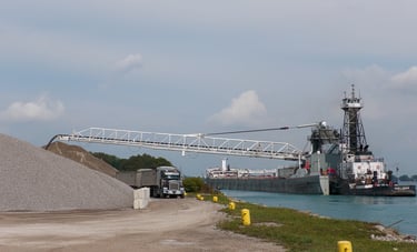 A semi-truck drives under the boom of a self-unloading barge, unloading at a stone yard.