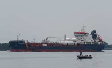 A man is standing up fishing in a small boat as a tanker ship passes behind him.