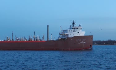 Front section of a classic forward pilothouse red and white freighter passing an oil refinery.
