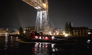 A Classic red and green tugboat in the dark, passing under the lit-up Duluth Aerial Lift Bridge.