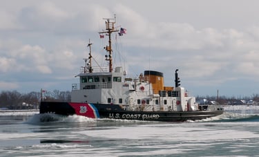 A Black and white U.S. Coast Guard tugboat making a large wake on icy water.