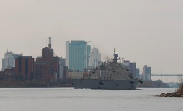 Right-side stern view of a gray navy ship passing some high-rise buildings.