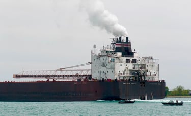 The aft section of a huge ship with a plume of steam exhaust, passing 2 small boats.