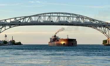 Sun shines on the bow of a huge red freighter, with a steam exhaust plume, passing under a bridge.