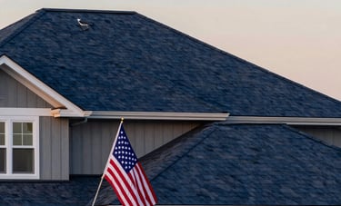 A reliable and sturdy home exterior in North American / US style with a newly installed dark blue roof. The lighting is the golden hour of sunset, highlighting the textures of the roof. In the yard, an American flag is neatly displayed on a pole, conveying patriotic pride. North American / US.