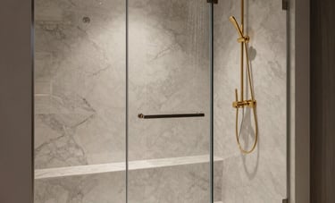 Interior photography of a luxury walk-in shower. Features include floor-to-ceiling porcelain tiles, a built-in bench, a ceiling-mounted rainfall showerhead in gold, and a seamless glass door. The style is modern and high-end.