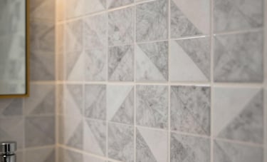 Close-up photography of a beautifully finished bathroom featuring intricate geometric tile patterns in light gray and white. Soft gold accent lighting illuminates the wall, highlighting the high-quality craftsmanship and modern fixtures.