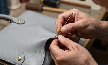 Detail of a master artisan's hands performing delicate stitching on a designer leather handbag. The workshop setting is professional and timeless, featuring a palette of soft grey and antique gold. High-end photography style focusing on the precision of the restoration process in a Portuguese atelier.