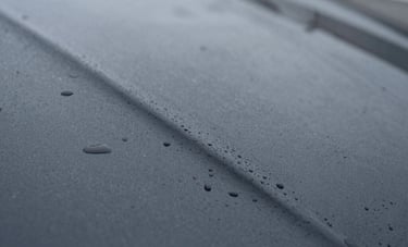 A macro, detailed photograph of a smooth, dark gray road surface in a Northern European setting. Water droplets are beading on the waterproof seal of a fresh repair.