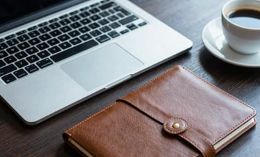 Close-up photography of a professional workplace in Western European / French style. A silver laptop, a leather-bound notebook, and a cup of coffee sit on a dark wood desk. The lighting uses navy and sky blue tones to create a sense of focused expertise.