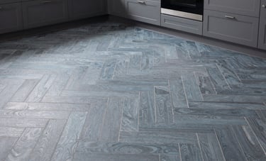 Wide shot of a modern North American kitchen featuring a stunning herringbone tile floor in muted blue-grey. The lighting is natural and soft, showcasing the precision of the installation and the sophisticated atmosphere of the home.