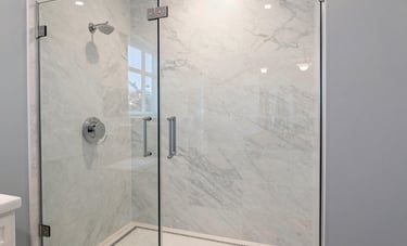 A luxurious bathroom remodel in a North American home, featuring a marble-tiled walk-in shower with a sleek glass door. The color palette includes off-white and pale blue accents, conveying a sense of high-end craftsmanship and modern elegance.