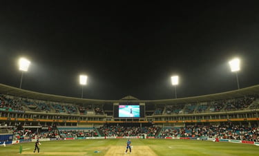 Cinematic wide-angle photography of a majestic South Asian cricket stadium at night, bright golden floodlights illuminating the pitch, dark sky, capturing high-energy atmosphere.