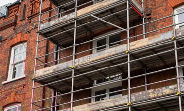 A professional shot of a high-quality scaffolding installation surrounding a traditional London red-brick building. Sharp focus on safety rails and yellow footings, bright daylight, Northern European / British architecture.