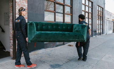 Professional movers carrying a green velvet sofa out of a building on a city sidewalk.
