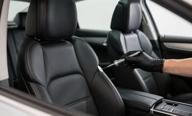 Interior detailing of a premium European sedan featuring black leather seats and silver accents. A professional hand in a black glove uses a precision tool to clean the stitching. The scene is bright and minimalist, shot with a shallow depth of field in a Western European detailing studio.