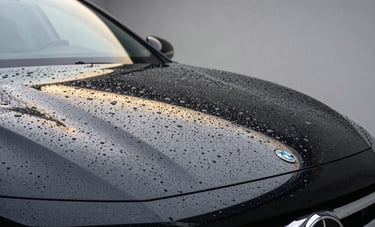 A close-up shot of water beading perfectly on a black car hood treated with ceramic coating. The surface is like a mirror, reflecting a modern studio with gold and silver light tones. The composition is artistic and minimalist, embodying extreme precision and automotive luxury.
