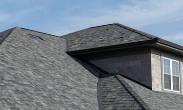 A low-angle, wide-shot photograph of a newly installed, modern roof on a large suburban home in North America, featuring clean lines and high-quality dark gray architectural shingles under a blue sky.