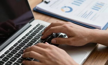 Close-up of hands typing on a modern laptop in a professional Brazilian workspace. Next to the laptop, a neatly prepared document with navy blue accents and professional charts. Shallow depth of field, focused on efficiency and detail.